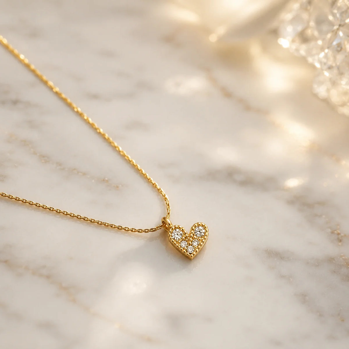 A delicate gold chain necklace with a small heart-shaped pendant paved with clear stones, resting on a white marble surface with warm, soft-focus light reflections.