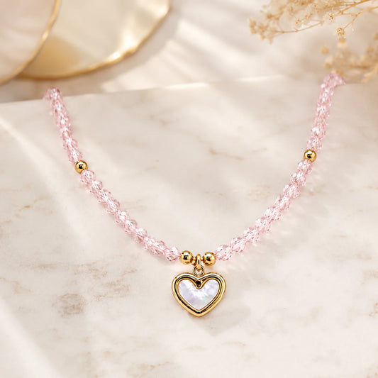 A delicate pink beaded necklace with a gold heart-shaped pendant featuring a white mother-of-pearl center, displayed on a light marble surface.