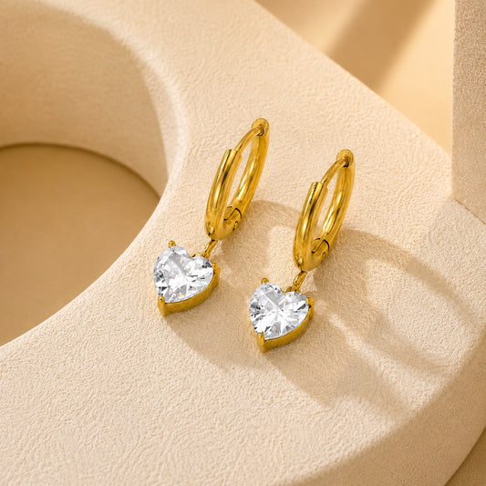 A pair of gold hoop earrings with heart-shaped clear crystal drops, displayed on a textured beige jewelry stand.