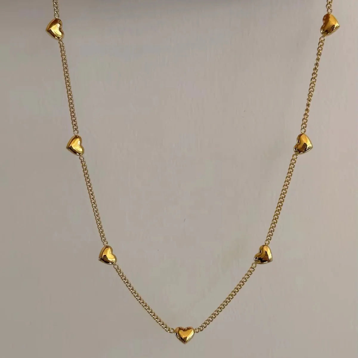 A gold chain necklace featuring multiple small, polished heart-shaped charms spaced along the chain, displayed against a plain light gray background.